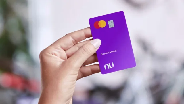 ão segurando um cartão de crédito roxo do Nubank com o nome "Suzana Amaral", exibindo o logotipo "Nu" e a bandeira Mastercard, ilustrando os resultados do Nubank 4T25.