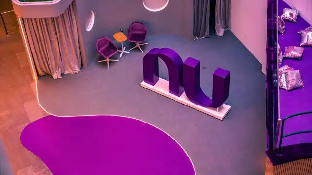 Vista superior do lounge da sede do Nubank, apresentando um grande logotipo "nu" roxo esculpido no centro, poltronas de design moderno e áreas de convivência em tons de violeta, representando a Nubank BBI.