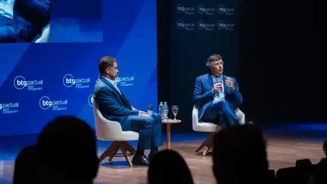 André Esteves e Mansueto Almeida sentados em poltronas brancas em um palco com painéis azuis da BTG Pactual Asset Management, participando de um debate sobre investimentos.
