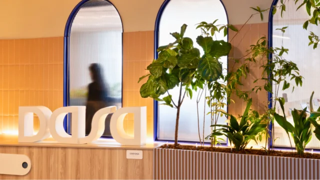 Logotipo tridimensional branco da Dasa posicionado sobre um balcão de madeira em uma recepção iluminada com plantas decorativas, representando a identidade da dasa dasa3.