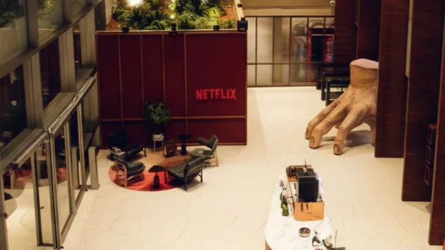 Vista aérea do saguão da sede da Netflix, apresentando uma área de estar com poltronas modernas, uma parede vermelha com o logotipo da marca e uma escultura gigante da "Mãozinha" (da série Wandinha) no chão, ilustrando a Netflix Warner.