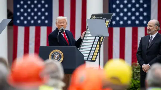 O presidente Donald Trump em um púlpito oficial, segurando um painel com uma lista de tarifas recíprocas ("Reciprocal Tariffs") diante de bandeiras dos Estados Unidos.