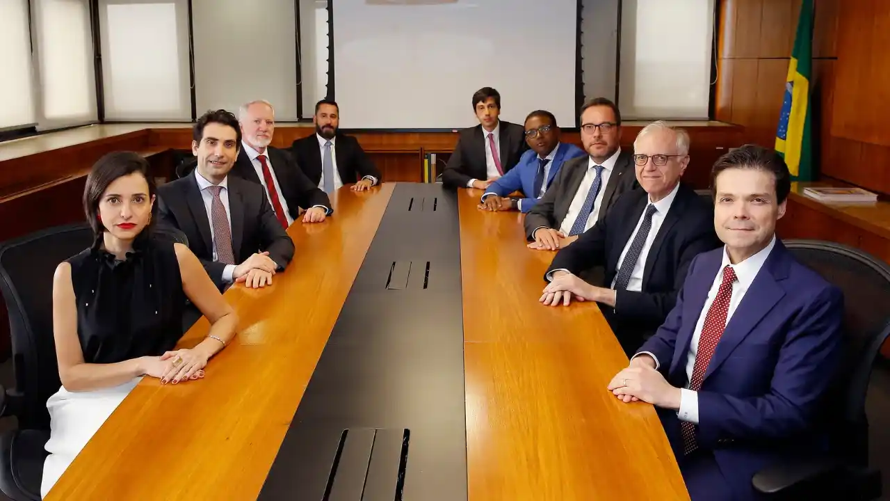 Fileira de microfones pretos de conferência posicionados sobre uma bancada em um auditório corporativo com painéis de madeira ao fundo. Em primeiro plano e ligeiramente desfocado, um bloco de notas com o logotipo azul do Banco Central do Brasil repousa ao lado de uma caneta.