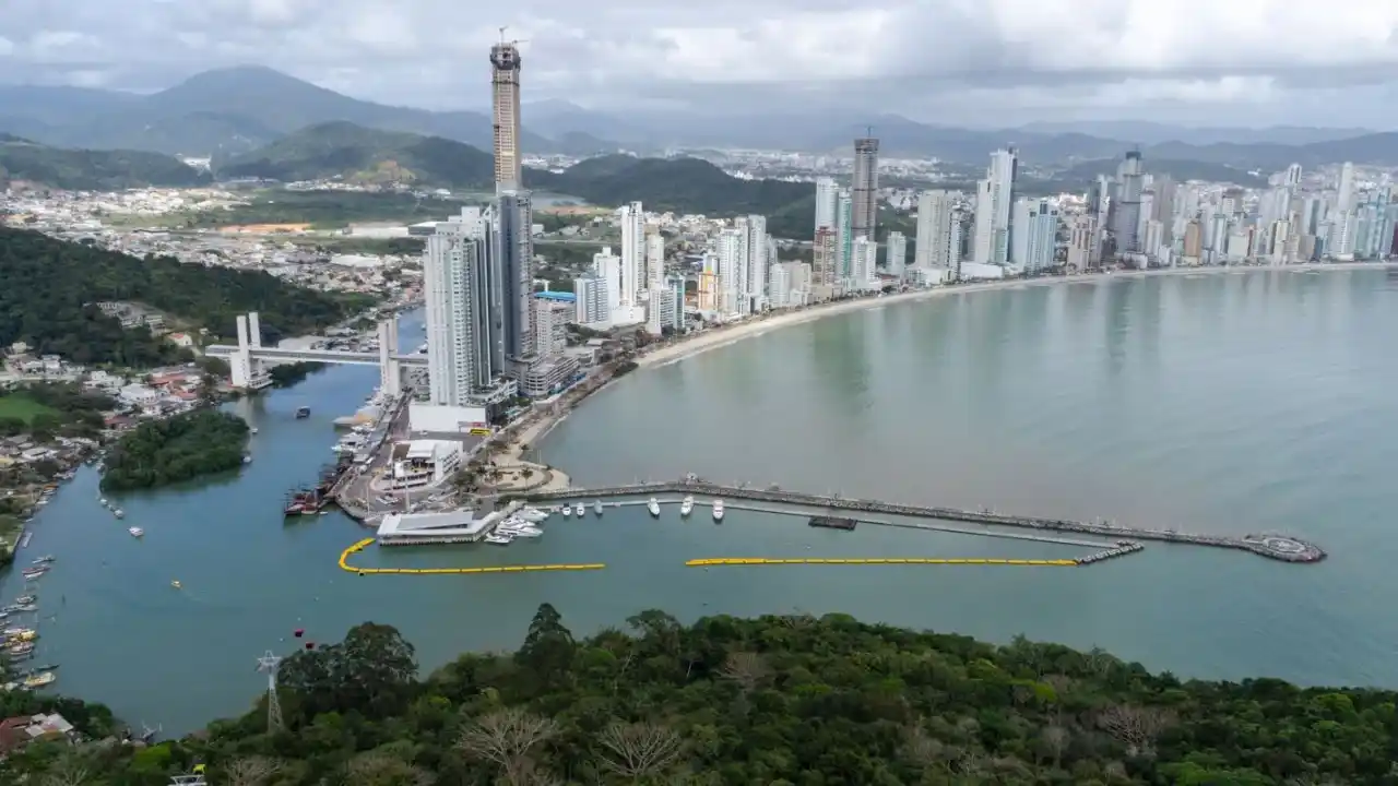 Vista aérea panorâmica da orla de Balneário Camboriú, Santa Catarina, destacando o mar, a faixa de areia, os grandes arranha-céus da Barra Sul e o molhe com barcos atracados no Rio Camboriú