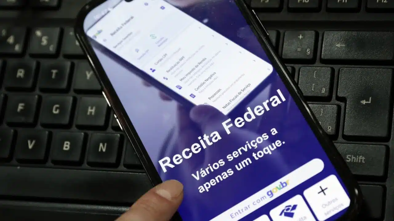 Close-up de uma mão segurando um smartphone sobre um teclado de computador. A tela do celular exibe o aplicativo da Receita Federal com a mensagem "Vários serviços a apenas um toque" e o botão de login "Entrar com gov.br". Opções como "Meu Imposto de Renda" e "Restituição IRPF" são visíveis na interface.
