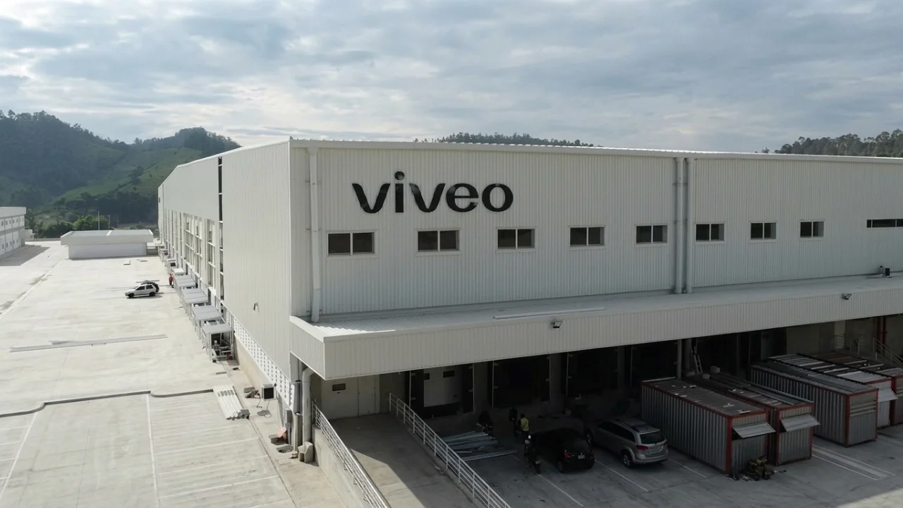 Vista aérea lateral de um grande centro de distribuição da Viveo. O galpão industrial é de cor branca com o logotipo "viveo" em letras pretas na fachada. A estrutura possui diversas docas de carga com caminhões e contêineres estacionados, um pátio pavimentado amplo e está cercada por colinas verdes ao fundo sob um céu nublado.