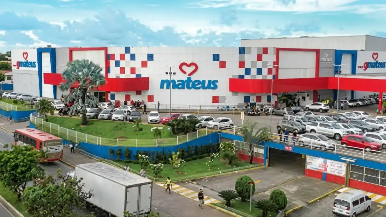 Vista aérea e panorâmica em ângulo diagonal de um grande e moderno supermercado do Grupo Mateus durante o dia. A fachada do edifício é predominantemente branca com detalhes geométricos em formato de xadrez nas cores azul e vermelho, exibindo ao centro o logotipo oficial composto por um coração vermelho vazado e a palavra "mateus" em letras azuis. Em frente à loja, há um amplo estacionamento ao ar livre completamente preenchido por carros e picapes de diversas cores. No primeiro plano inferior, vê-se uma avenida urbana com um ônibus vermelho de transporte público, um caminhão baú branco, pedestres caminhando por uma faixa de pedestres amarela e um canteiro ajardinado com uma palmeira imponente. O céu ao fundo está parcialmente nublado.