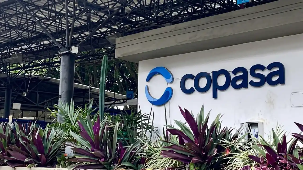 Fotografia em plano médio da entrada de uma instalação da Copasa. Fixado em uma parede branca texturizada, destaca-se o logotipo azul da companhia — formado por duas formas curvas estilizadas que se completam — seguido pelo nome "copasa" em letras minúsculas azuis. Em primeiro plano, canteiros densos com plantas tropicais ornamentais de folhas roxas e verdes decoram a base da estrutura. Ao fundo e na parte superior, vê-se uma cobertura industrial com treliças metálicas pretas sob a copa de árvores.