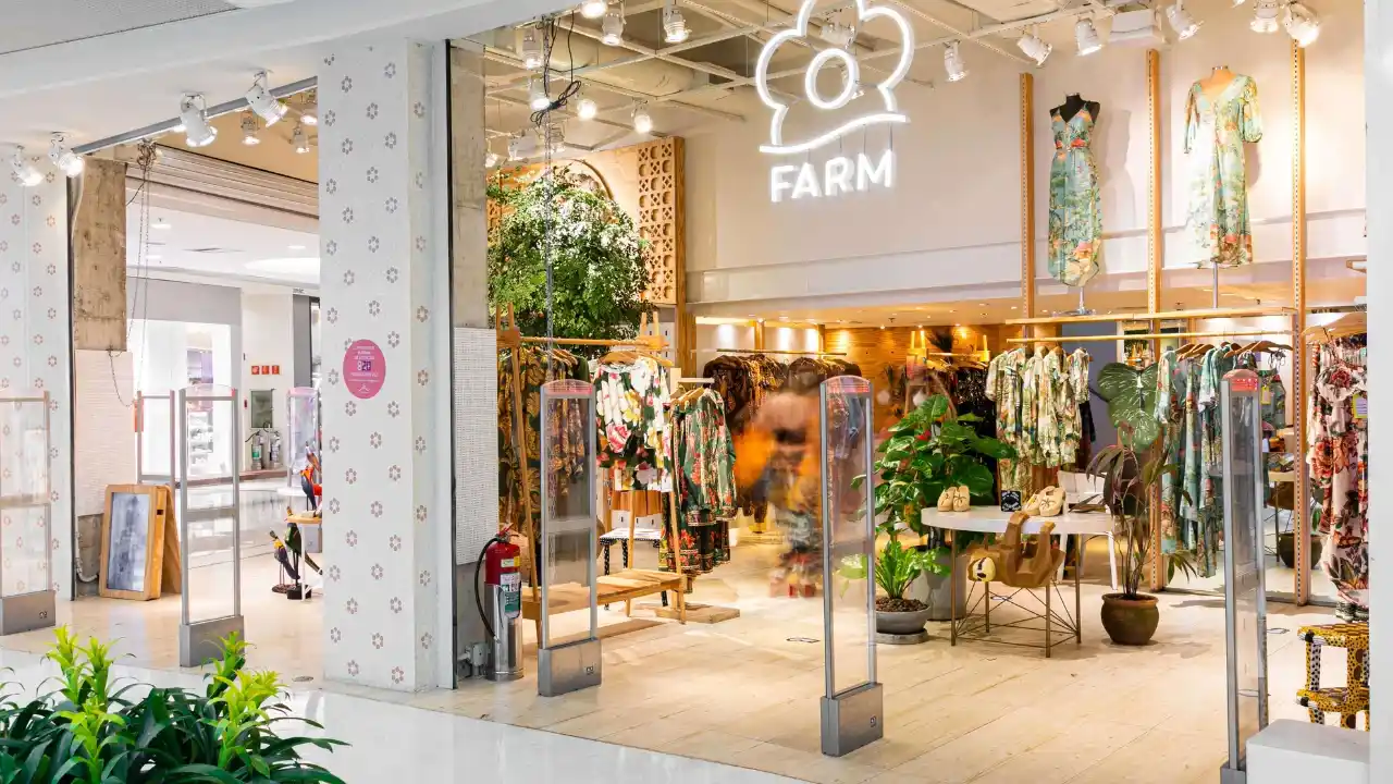 Fotografia em plano aberto da entrada de uma loja da Farm no Shopping Leblon. No topo da fachada interior branca, destaca-se o logotipo iluminado em neon com o desenho de uma flor estilizada e a palavra "FARM" logo abaixo. O interior do estabelecimento apresenta piso e painéis de madeira clara, araras repletas de roupas com estampas coloridas e tropicais, manequins exibindo vestidos longos em suportes elevados, plantas de folhas verdes grandes em vasos de cerâmica e mesas centrais com calçados. Na entrada, há dois totens de segurança transparentes e um canteiro com folhagens verdes em destaque no canto inferior esquerdo sob iluminação spot focada.