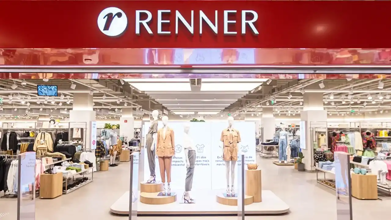 Fotografia em plano aberto da entrada de uma loja física da Lojas Renner. Na parte superior, destaca-se um grande painel horizontal vermelho brilhante com o logotipo da marca — a letra "r" minúscula estilizada em branco dentro de um círculo — ao lado do nome "RENNER" em letras maiúsculas brancas e iluminadas. O interior da loja é amplo, moderno e muito bem iluminado por painéis de LED no teto, exibindo manequins vestidos com roupas de alfaiataria em tons pastéis e neutros sobre plataformas circulares, além de araras de roupas organizadas e displays promocionais ao fundo.