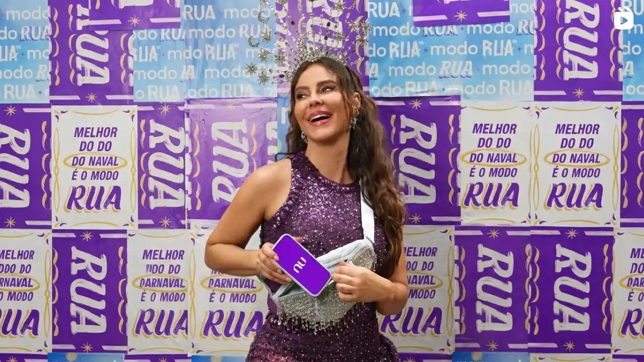 Fotografia em plano médio de uma mulher sorrindo, vestindo um vestido roxo de paetês brilhantes e uma tiara prateada detalhada na cabeça. Ela segura e guarda um smartphone com capinha roxa estampada com o logotipo branco "nu" dentro de uma pequena bolsa transversal prateada e brilhante. O fundo é uma parede preenchida por cartazes lambe-lambe repetitivos nas cores roxo, branco e amarelo com os textos "modo RUA" e "MELHOR DO CARNAVAL É O MODO RUA".
