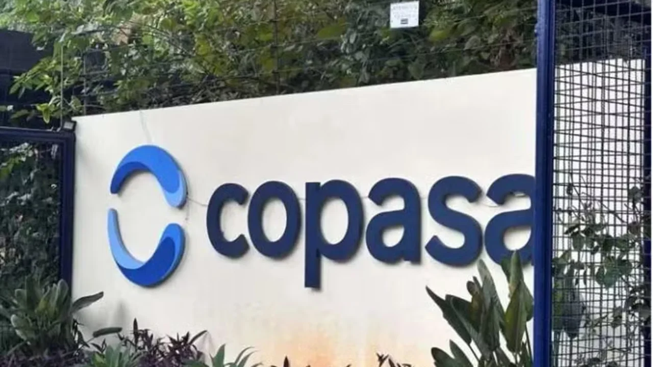 Fotografia em plano médio da fachada institucional externa da Copasa sob a luz natural do dia. Em uma parede de alvenaria branca e lisa ao centro, destaca-se à esquerda o logotipo circular estilizado composto por arcos em tons de azul e branco, seguido pela palavra "copasa" escrita em letras minúsculas e relevo azul-escuro. Na base da parede, há pequenas plantas rasteiras e folhagens tropicais escuras. As laterais da estrutura são delimitadas por grades e portões de ferro pintados em azul-escuro, enquanto árvores e vegetação densa cobrem o fundo sob um céu claro.