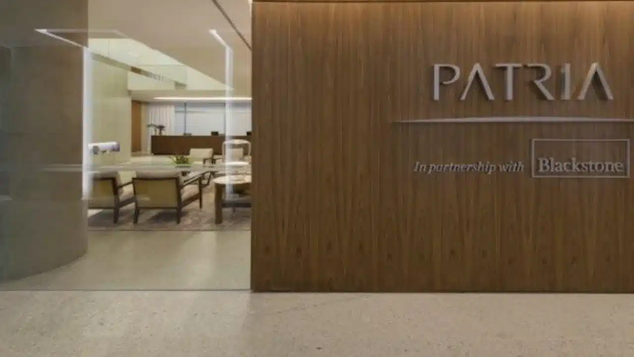 Entrada da sede da Pátria Investimentos com elegante parede revestida em painéis verticais de madeira maciça. Ao centro da parede, sobressai a palavra "PATRIA" em letras prateadas tridimensionais com design estilizado, acompanhada do texto "In partnership with Blackstone" em moldura ao lado. À esquerda, através de uma divisória de vidro transparente, avista-se a recepção com poltronas beges, iluminação embutida e piso de mármore claro.