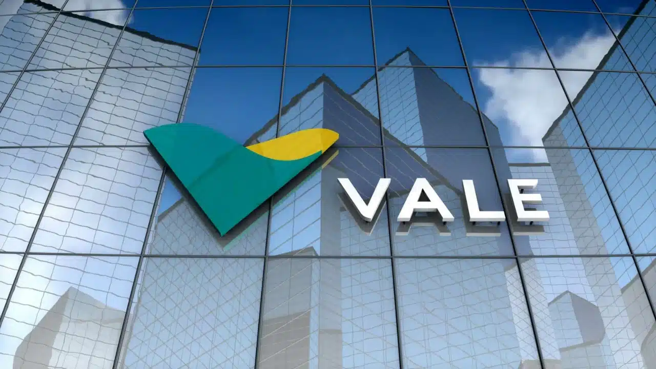 Fachada em ângulo inferior de um prédio corporativo moderno com placas de vidro espelhado refletindo o céu azul com nuvens. No centro do painel de vidro, destaca-se a logomarca tridimensional da Vale, formada pelo símbolo estilizado em "V" nas cores verde e amarela ao lado da palavra "VALE" escrita em letras maiúsculas na cor branca.