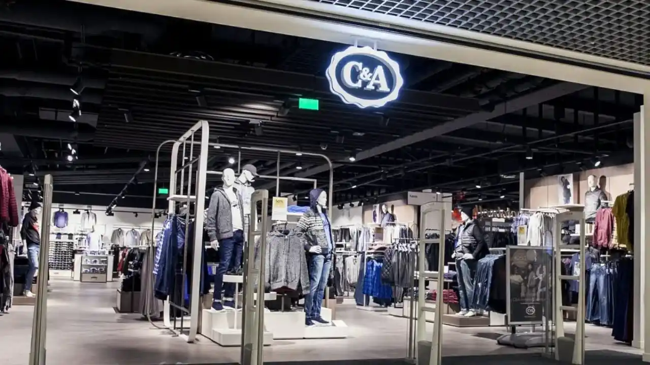 Entrada de uma loja da C&A em shopping center, com o logo luminoso da marca em destaque no teto, sensores antifurto nas laterais e manequins ao centro vestindo jaquetas, moletons e calças jeans, cercados por araras de vestuário.