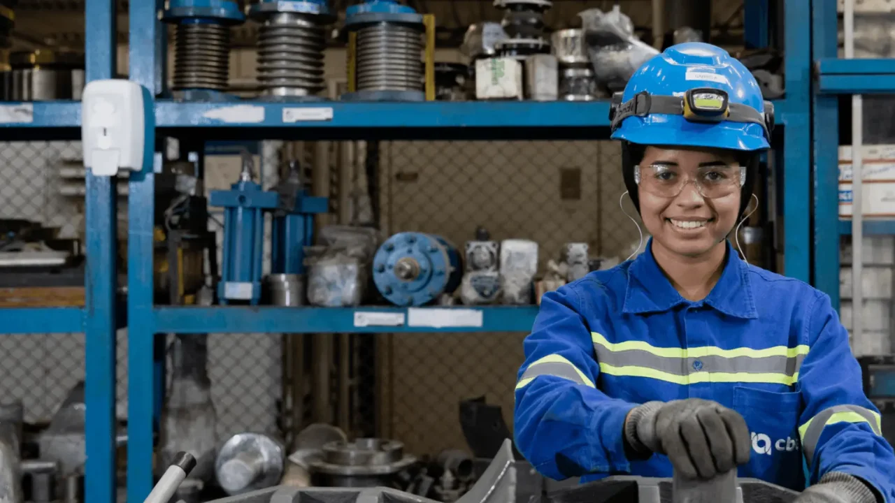 Trabalhadora industrial sorrindo em uma oficina de manutenção da CBA, vestindo uniforme azul de proteção com o logotipo impresso na manga, capacete azul com lanterna de cabeça e óculos de segurança. Ao fundo, prateleiras organizadas contendo peças metálicas, cilindros hidráulicos e equipamentos industriais.