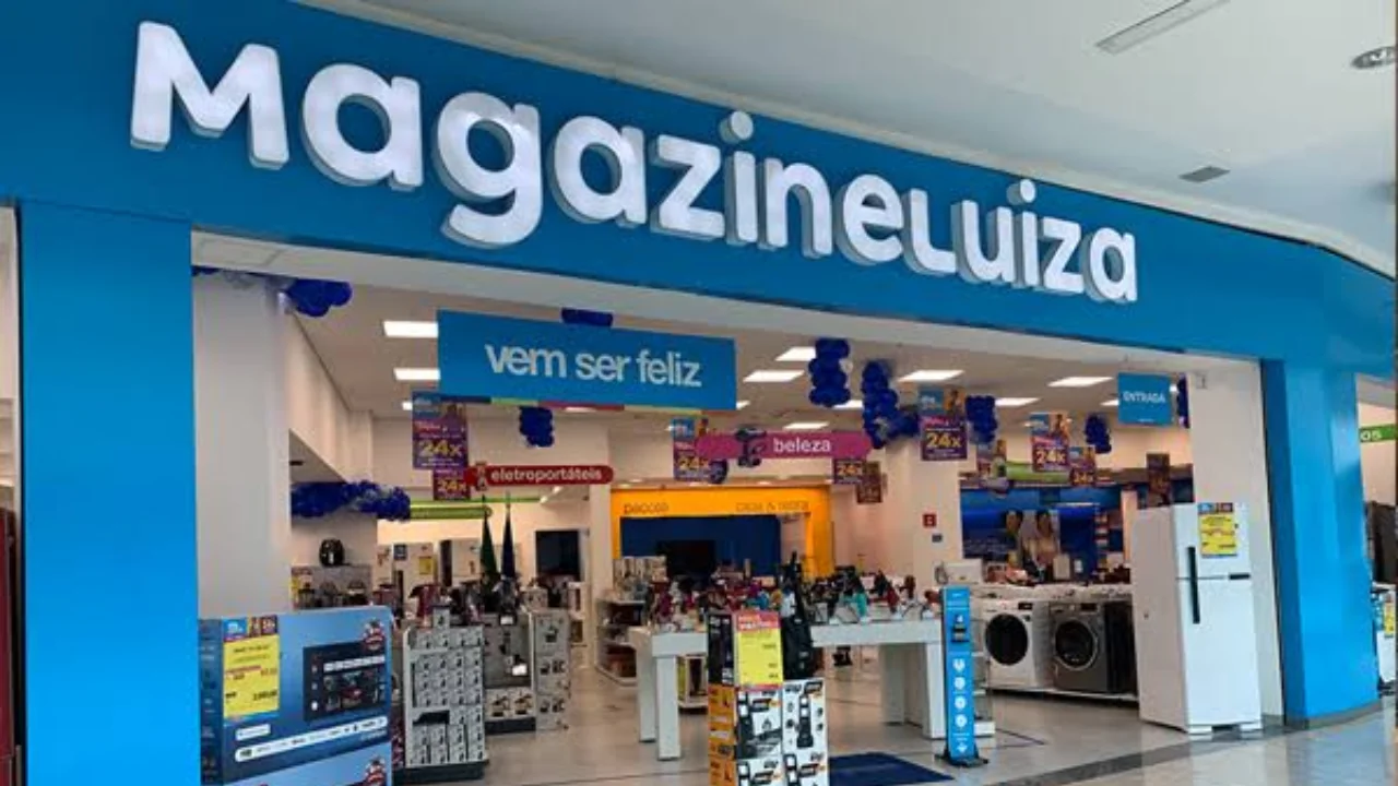 Fachada ampla de loja física do Magazine Luiza em shopping center com letreiro luminoso azul, cartaz central com a frase "vem ser feliz" e ambiente interno exibindo refrigeradores, televisores e eletroportáteis.