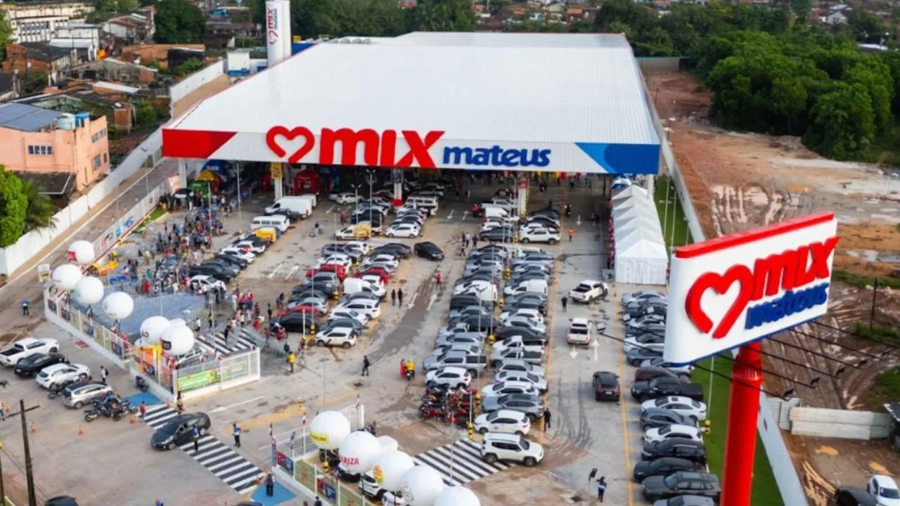 Vista aérea de loja do Mix Mateus com fachada branca e vermelha, totem com logotipo da marca, estacionamento coberto e descoberto com carros e balões promocionais de inauguração.