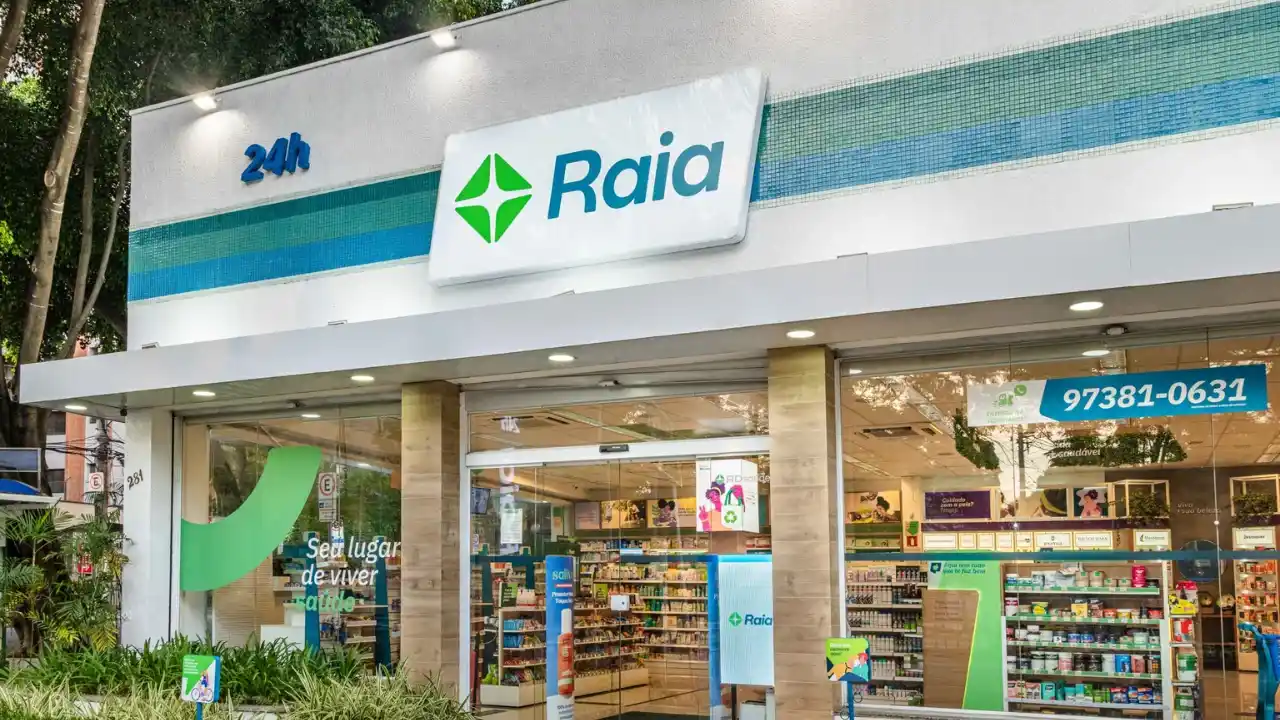 Fachada externa e iluminada de uma farmácia Droga Raia, bandeira da RD Saúde, com estrutura branca moderna, letreiro azul e verde com o logo da marca, indicação de funcionamento 24 horas e grandes vitrines de vidro exibindo o interior organizado da loja.