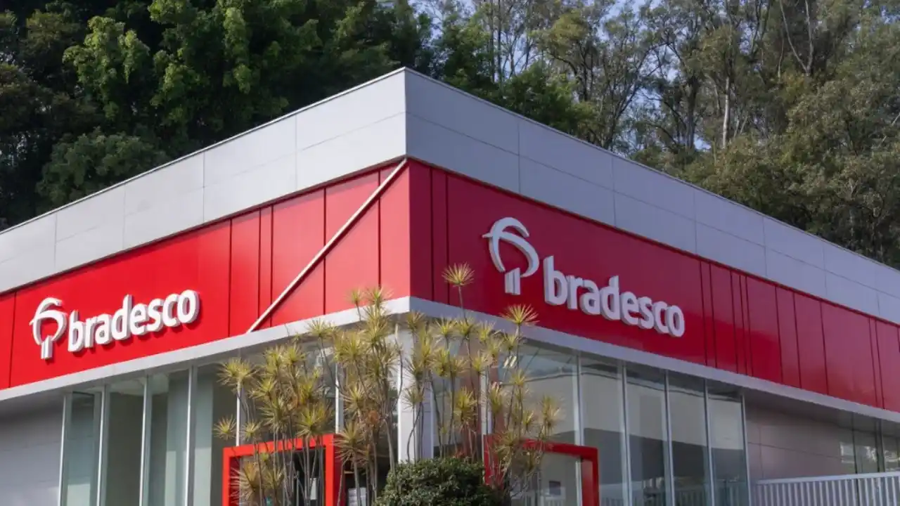 Fachada moderna de uma agência bancária do Bradesco com estrutura vermelha e branca, portas de vidro e o logotipo do banco em destaque, cercada por vegetação e árvores ao fundo.