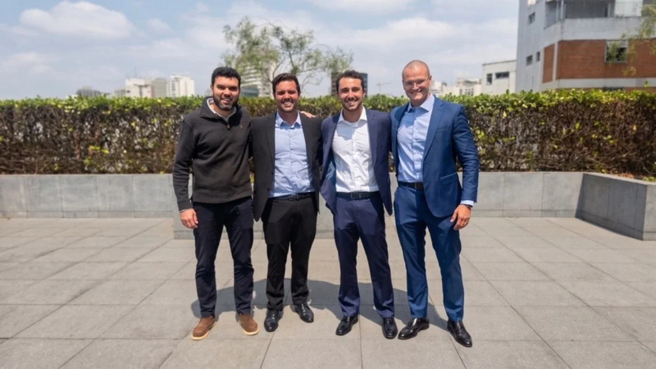 Quatro executivos sócios vestindo trajes esporte fino e ternos alinhados lado a lado em uma área externa ao ar livre com jardim e prédios ao fundo.
