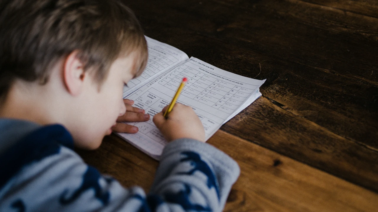 Criança escrevendo em uma folha de atividades escolares com lápis sobre uma mesa de madeira.