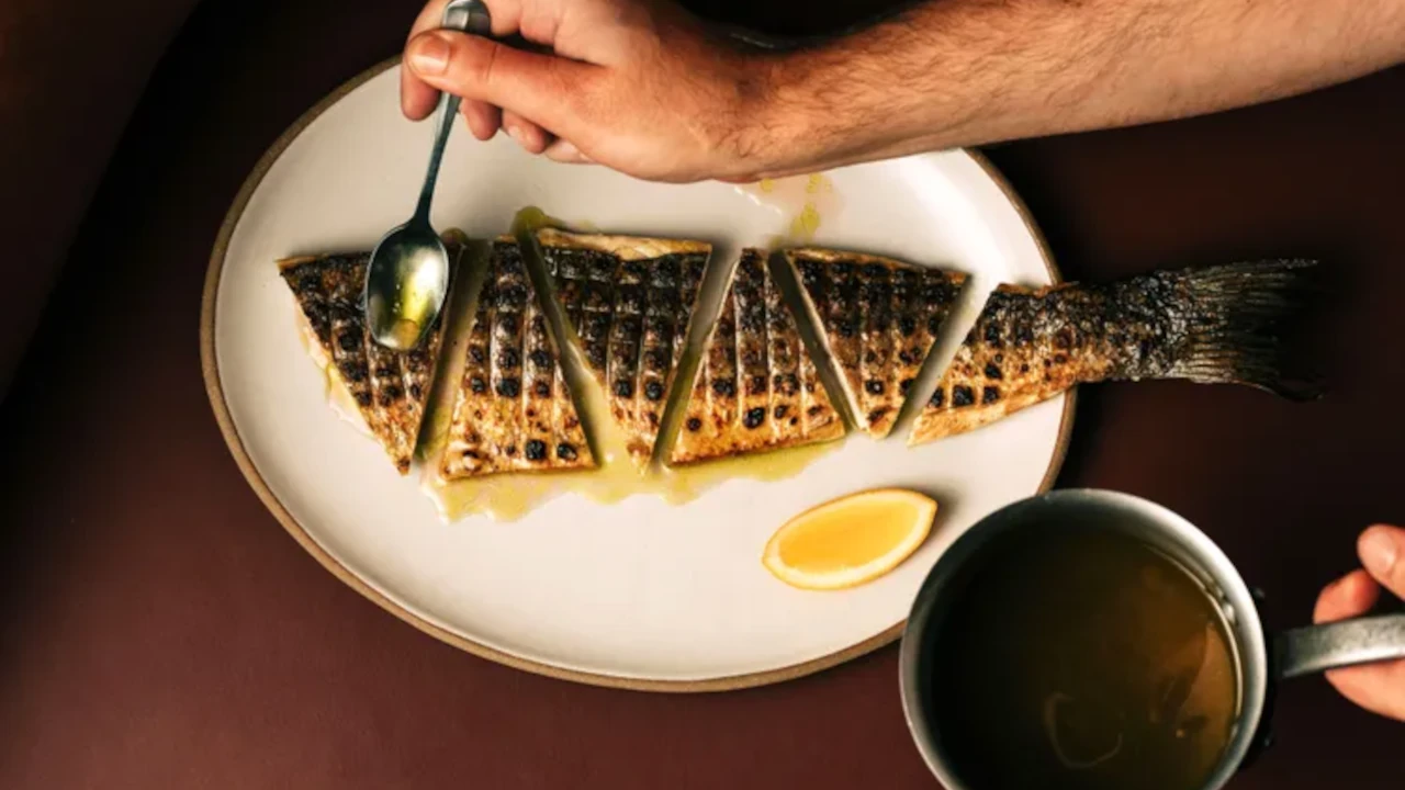 Robalo inteiro grelhado e servido em fatias sobre prato branco, finalizado com molho e acompanhado por limão, em restaurante de Daniel Humm.
