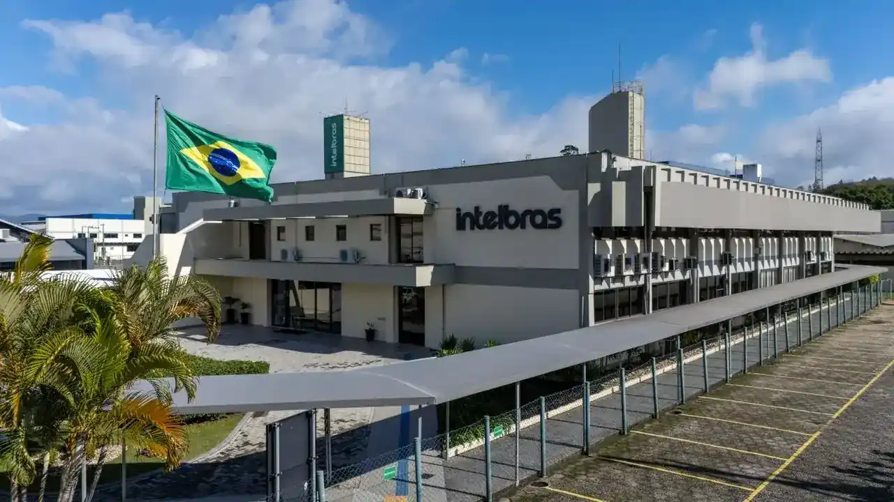 Fachada da sede da Intelbras em São José (SC), com edifício corporativo cinza, letreiro com a marca em destaque, bandeira do Brasil hasteada ao lado de palmeiras e caminho coberto na entrada sob céu ensolarado.