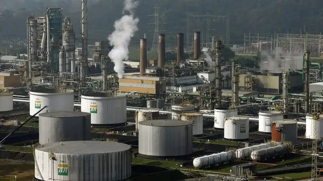 Vista aérea industrial de grandes tanques de armazenamento cilíndricos brancos e cinzas estampados com a logomarca da Petrobras, torres de refinamento e chaminés expelindo fumaça na Refinaria Presidente Bernardes em Cubatão.