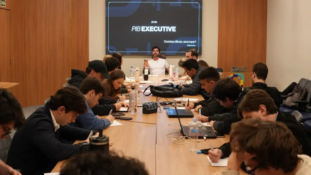 Estudantes sentados ao redor de uma grande mesa de reunião em sala de aula moderna, fazendo anotações e utilizando notebooks, enquanto um apresentador ao fundo ministra uma aula com a tela "PIB Executive" em destaque.